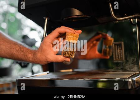 Abgeschnittene Ansicht des Barista, der Kaffeebohnen in einem Glas in der Nähe einer unscharfen Kaffeemaschine im Café hält Stockfoto