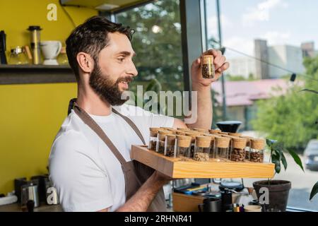 Positiver bärtiger Barista hält Gläser mit Kaffeebohnen, während er im Coffee Shop arbeitet Stockfoto