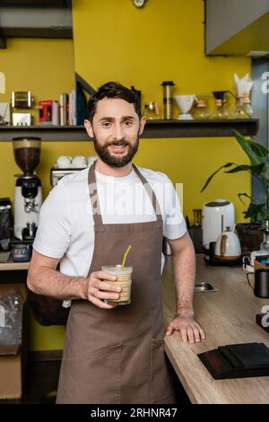 Positiver Barista in der Schürze, der ein kaltes Kaffeegetränk im Glas hält und im Coffee Shop vor der Kamera lächelt Stockfoto