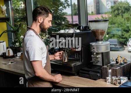 Seitenansicht des bärtigen Barista, der beim Arbeiten mit der Kaffeemaschine im Coffee Shop Milch aufschäumt Stockfoto