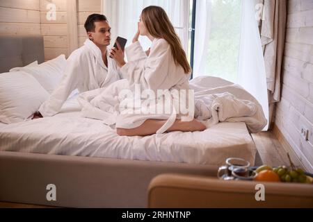 Mann und Frau tragen Bademäntel und sitzen auf weißen Laken im Bett Stockfoto