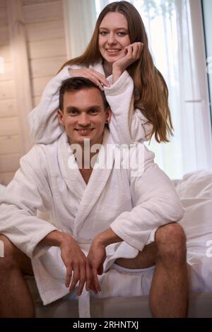 Glückliches Paar in weißen Bademänteln, die auf dem Bett sitzen Stockfoto