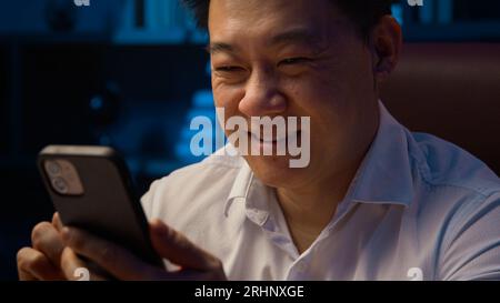 Glücklicher lächelnder asiatischer Geschäftsmann mittleren Alters koreanischer erwachsener Geschäftsmann chinesischer Arbeiter japanischer Arbeitgeber am Abend Home Office, das Handy durchsucht Stockfoto