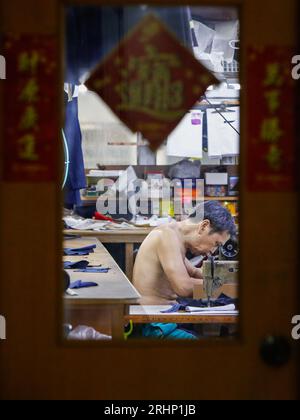 Hongkong, China. August 2023. Ein Schneider betreibt eine traditionelle Nähmaschine in einer Mini-Kleiderfabrik in Mirador Mansion, einem historischen Gebäude mit Wohnhäusern, erschwinglichen Pensionen und kleinen Unternehmen. Maßgeschneiderte Unternehmen dieser Art in Hongkong sind am Rande des Aussterbens, da sich die Konsumgewohnheiten im Laufe der Jahrzehnte verändert haben. Das Gebäude an der Nathan Road wurde 1959 gegründet, wo die Hersteller von Uhren oder Handschuhen und Schneidereien während der Industrialisierung zwischen den 50er und 70er Jahren blühten Aber jetzt gibt es nur noch ein paar Schneidereien und die meisten Stockfoto