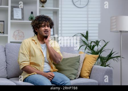 Junger kranker Mann sitzt zu Hause, hat starke Zahnschmerzen, hispanischer Mann auf Sofa im Wohnzimmer. Stockfoto