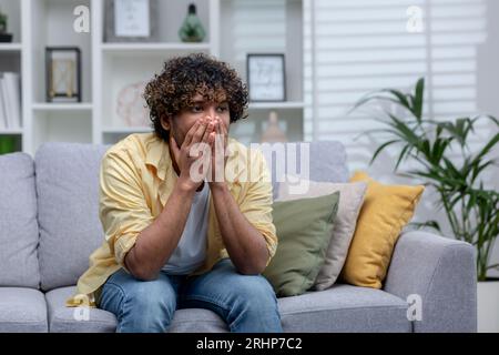 Trauriger depressiver Mann, der allein zu Hause auf dem Sofa im Wohnzimmer sitzt, verzweifelter indianer. Stockfoto