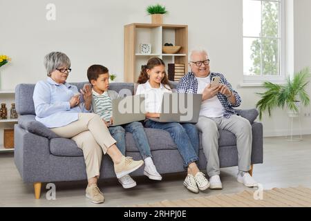 Glückliche Großeltern mit zwei Kindern, die zusammen mit Laptops und Mobiltelefonen auf dem Sofa sitzen. Stockfoto