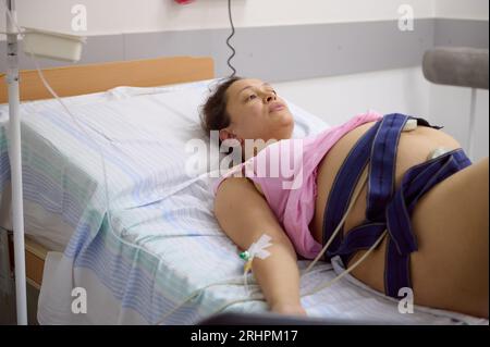 Schwangere Frau, die auf dem Bett in einer bequemen Station liegt, Kontraktionen hat, den Herzschlag und Puls ihres Babys überprüft, mit einem Elektrokardiogramm bel Stockfoto
