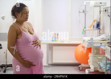 Multiethnische gebärende Mutter, die Kontraktion hat und ihren großen Bauch in der Geburtsstunde des Entbindungskrankenhauses hält. Schwangerschaft. Einfache Lieferung. Geburtshilfe A Stockfoto