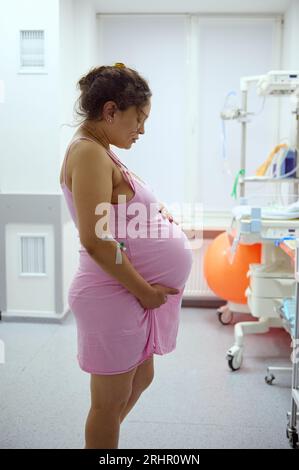 Seitenporträt einer schwangeren Frau, gebärende Mutter, die ihren Bauch während der Wehen hält, auf der Krankenhausstation. Schwangerschaft. Einfach Stockfoto