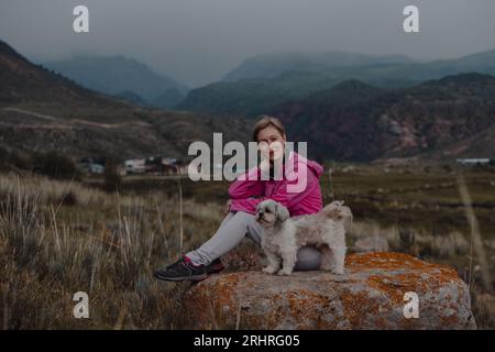 Junge Wanderin mit Shih-Tzu-Hund, die an einem bewölkten Tag auf einem Felsen in den Bergen sitzt Stockfoto