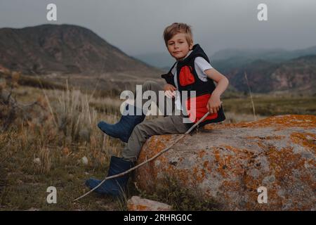 Junge Wanderer mit Stock in einer Weste und Gummistiefeln, die auf dem Stein in den Bergen sitzen Stockfoto