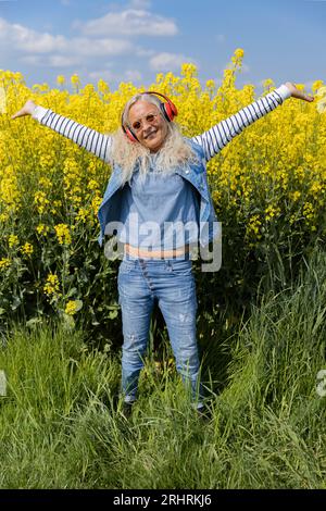 Fröhliche Seniorin mit Kopfhörern, die im Sommer unter einem Rapsfeld steht. Stockfoto