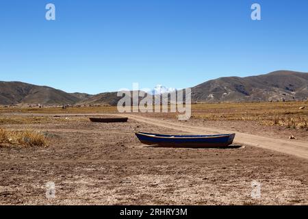 Titicacasee, BOLIVIEN; 18. August 2023: Hölzerne Ruderboote am ausgetrockneten Ufer der Cohana Bay am inneren See / Huiñay Marka (der kleinere Teil des Titicacasees) in der Nähe des Dorfes Cohana. Im Hintergrund befindet sich der Huayna Potosi (6088 m) in der Kordillera Real der Anden. Die Wasserstände im Titicacasee nähern sich dem Rekordtief von 1996, dem niedrigsten seit der Aufnahme von Aufzeichnungen durch den bolivianischen Wetterdienst (Senhami) im Jahr 1974. Viele geben dem Klimawandel die Schuld; die letzten Jahre waren trockener als normal, und El Niño verstärkt sich derzeit im Pazifischen Ozean vor der Südküste Stockfoto