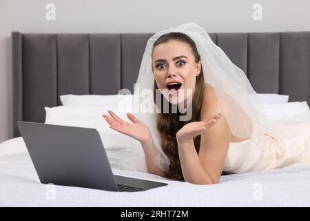 Schockierte Braut mit Laptop auf dem Bett im Schlafzimmer Stockfoto