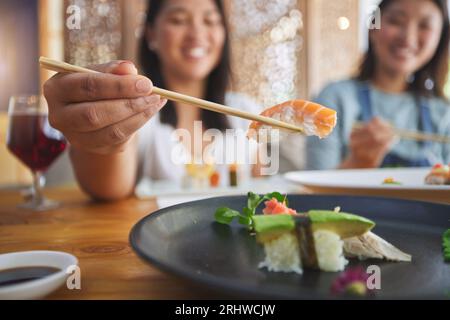 Japanische Frau, Essstäbchen und Nigiri im Restaurant, lächeln und freuen sich auf ein gutes Abendessen auf der Party. Asiatische Freunde, Fisch und gesund mit Kultur Stockfoto