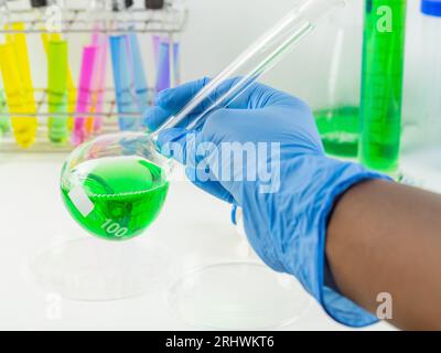 Nahaufnahme einer Hand mit Handschuhen, die einen Messkolben mit einer grünen Flüssigkeitsprobe in einem Labor hält. Stockfoto