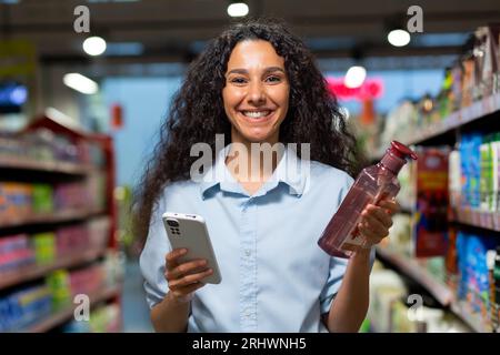 Porträt einer Käuferin in einem Supermarkt, eine hispanische Frau wählt Shampoo für Haare und Duschgel, liest Bewertungen im Internet, lächelt und verwendet eine Anwendung auf ihrem Telefon. Stockfoto