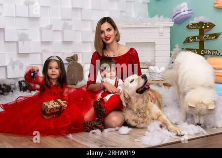 Familienfoto-Porträt. Mama und ihre zwei Kinder und zwei weiße Hunde in roten Kleidern feiern die Chistmas, Neujahr. Glückliche junge Mutter und ihre Tochter Stockfoto