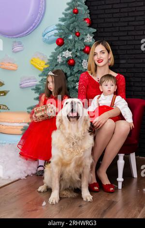 Familienfoto-Porträt. Mama und ihre zwei Kinder und ein weißer Hund in roten Kleidern feiern das neue Jahr der Chistmas. Glückliche junge Mutter und ihre Tochter, Stockfoto