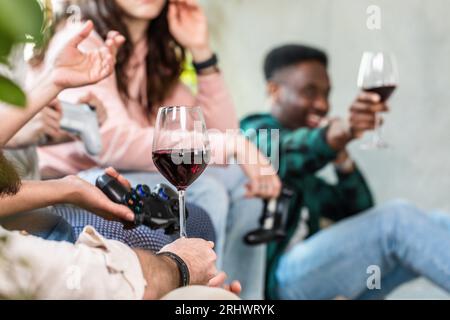 Selektiver Fokus auf die Hand eines Jungen, der einen Becher Rotwein hält. Multi-ethnische Partei unter Freunden mit nicht erkennbaren Menschen. Konsole für Spiele in einem han Stockfoto