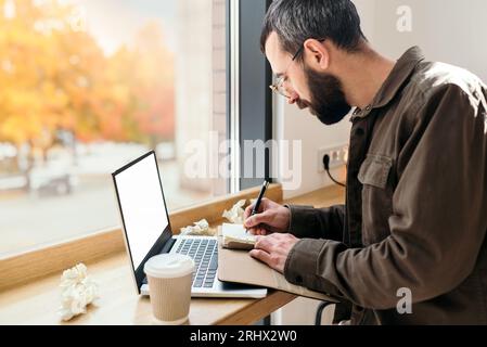 Bärtiger Geschäftsmann in lässigem Stil, der in einem Notizbuch in einem Café schreibt. Ein Mann sitzt in einem Café mit einem Laptop, Fernarbeit, Freiberufler, Herbst Stockfoto