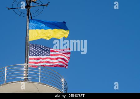 Die Flaggen der Ukraine und der USA flattern auf einem Fahnenmast vor einem blauen Himmel. Statussymbol. Ukrainische blaue und gelbe Flagge. Windtag im Freien. Stockfoto