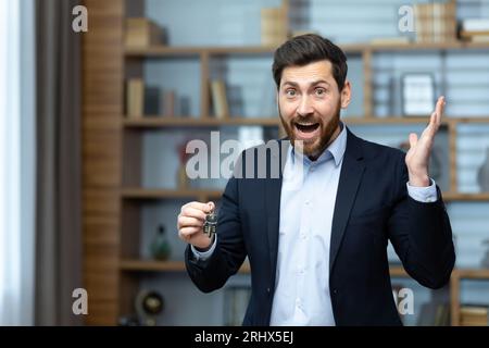 Porträt eines glücklichen und erfolgreichen Maklermaklers, der Mann schaut freudig auf die Kamera und hält die Schlüssel zu einer neuen Wohnung im Haus, ein Geschäftsmann in einem Business-Anzug arbeitet im Büro. Stockfoto