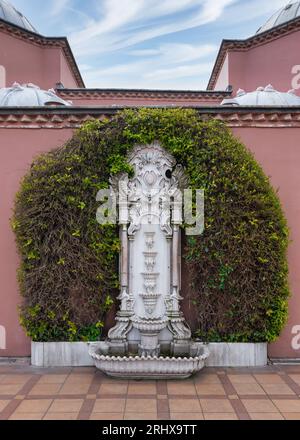 Weißer Marmorbrunnen am Sultan Ahmed Square, Istanbul, Türkei, umgeben von üppigem Grün und einer rosafarbenen Steinmauer des Hagia Sophia Hurrem Sultan Bathhouse oder Ayasofya Hurrem Sultan Hamami Stockfoto