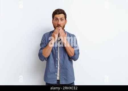 Gutaussehender Mann bedeckt seinen Mund mit seinen Händen mit schockiertem Ausdruck auf weißem Hintergrund Stockfoto