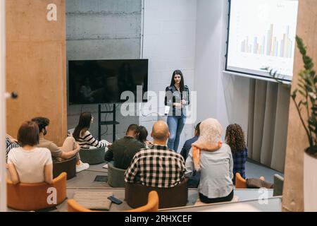 Junge und schöne Geschäftsfrau lächelt und präsentiert einer vielfältigen Gruppe von Menschen in einem kleinen Vorlesungssaal ein Diagramm Stockfoto