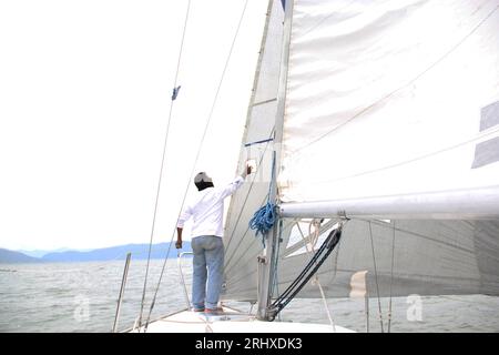 Ein Erwachsener Latino fährt ein Segelboot auf dem See mit Segeln, die als körperliche Aktivität, Sport und Hobby entwickelt wurden, um sich am Wochenende zu entspannen Stockfoto