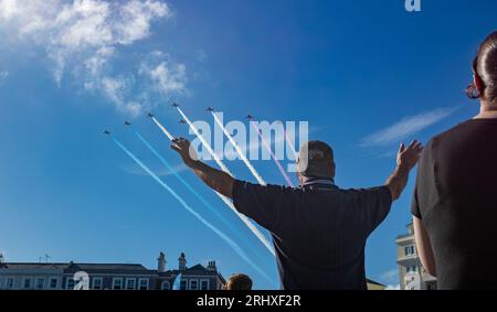 Eastbourne, Großbritannien. August 2023. Ein Mann am Strand von Eastbourne streckt seine Arme aus, während das weltberühmte RAF-Ausstellerteam The Red Arrows auf der jährlichen Eastbourne Airbourne, einer internationalen Flugshow, über ihm fliegt. Die Show findet vier Tage lang statt, mit Flugvorführungen entlang der Küste. Stockfoto