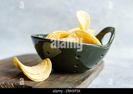 Gestapelte Stücke köstlicher knuspriger gelber Kartoffelchips in schwarzer Schüssel über Holztisch in hellem Raum vor verschwommenem Hintergrund Stockfoto