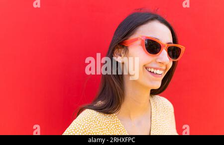Positive junge Frau in trendiger Sonnenbrille lächelt und schaut weg, während sie vor rotem Hintergrund steht Stockfoto