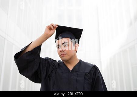 Porträt eines selbstbewussten, gutaussehenden jungen Mannes, der ein schwarzes Abschlusskleid trägt, das die Kappe berührt, die mit gegen die weiße Wand steht Stockfoto