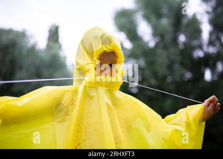 Nicht erkennbares Kind in gelbem Regenmantel, das bei regnerischem Wetter tagsüber vor unscharfen grünen Bäumen im Wald steht Stockfoto