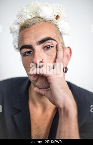 Porträt eines jungen, unrasierten Mannes mit kurzen Haaren, blauen Schnurrbart-Augen und Blumenkrone, die in die Kamera schauen, während sie die Hand auf das Gesicht in einem verschwommenen Raum zurücklehnen Stockfoto