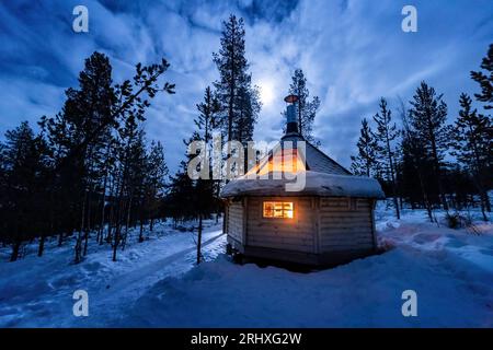 Außenseite einer kleinen beleuchteten Holzhütte, die sich im Winter nachts in schneebedeckten Wäldern in Lappland, Norwegen, befindet Stockfoto
