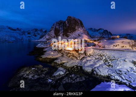 Spektakulärer Blick auf beleuchtete kleine Häuser an der verschneiten Küste gegen felsige Berge auf den Lofoten-Inseln in Norwegen bei Nacht Stockfoto
