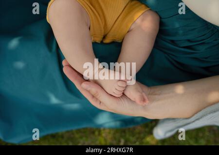 Hochwinkelige, abgeschnittene Hand einer unerkennbaren kaukasischen Mutter, die die Beine des Babys hält, während sie im Sommer am Wochenende im Park sitzt Stockfoto