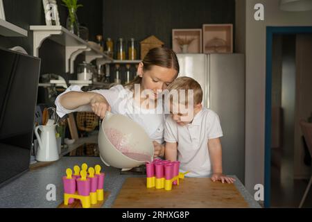 Kaukasischer Sohn mit blondem Haar, der neugierig auf die Mutter schaut, die Flüssigkeit in Eiswürfelformen gießt, während sie in der Küche in der Freizeit Eis zubereitet Stockfoto
