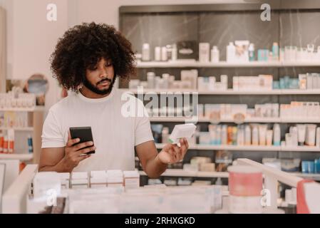 Afro-amerikanischer Mann mit Afro-Frisur, der Smartphone hält und Informationen auf der verschriebenen Arzneimittelverpackung liest, während er mit gegen die Regale steht Stockfoto