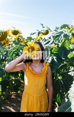 Lächelndes kaukasisches junges Mädchen, gekleidet in gelben Casuals, das Gesicht mit wunderschöner Sonnenblume versteckt, während es inmitten von Pflanzen auf dem Bauernhof steht Stockfoto