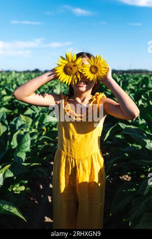 Lächelndes kaukasisches junges Mädchen, gekleidet in gelben Casuals, das Gesicht mit wunderschönen Sonnenblumen versteckt, während es inmitten von Pflanzen auf dem Bauernhof steht Stockfoto