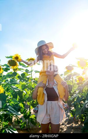 Kaukasische mittelErwachsene Mutter, die lächelnde, entzückende Tochter auf den Schultern trägt, während sie im Sonnenblumenfeld gegen klare s steht und sich anschaut Stockfoto