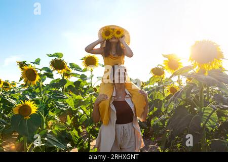 Kaukasische mittelErwachsene Mutter, die auf Schultern trägt verspielte Tochter, die im Sommer Sonnenblumen auf dem Bauernhof vor klarem Himmel hält Stockfoto