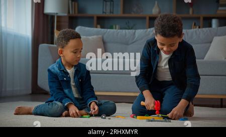 Zwei afroamerikanische Brüder, kleine ethnische Jungen, Kinder, Geschwister, Freunde, Kinder im Wohnzimmer, die mit Spielzeugen spielen, die das Gebäude reparieren Stockfoto