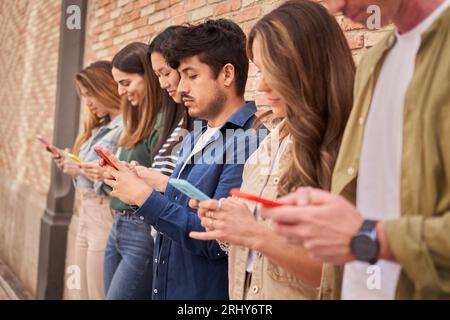 Seitenansicht multirassische Gruppe konzentrierte junge Generation z lehnte Ziegelwand mit Telefonen im Freien. Stockfoto