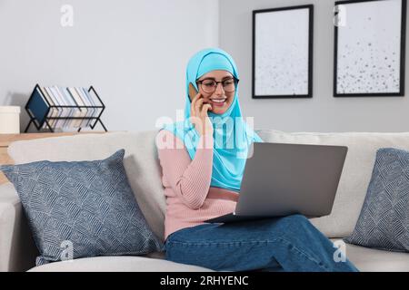 Muslimische Frau, die auf dem Smartphone in der Nähe des Laptops auf der Couch im Zimmer spricht Stockfoto